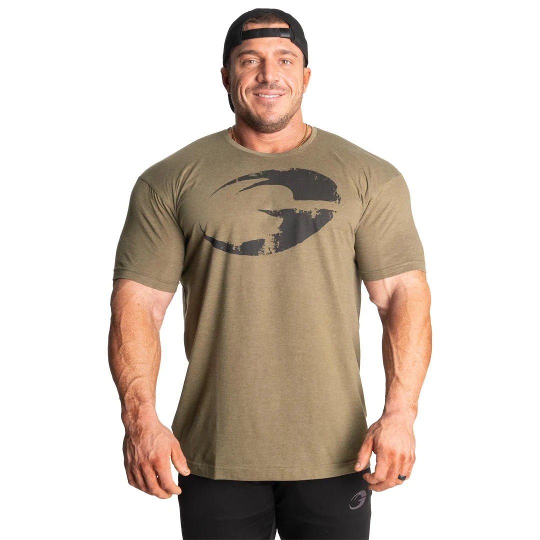 GASP Cadet Tee Army Green Melange, en grönmelerad t-shirt med svart logga på bröstet buren av en leende man., bild 6 av 8