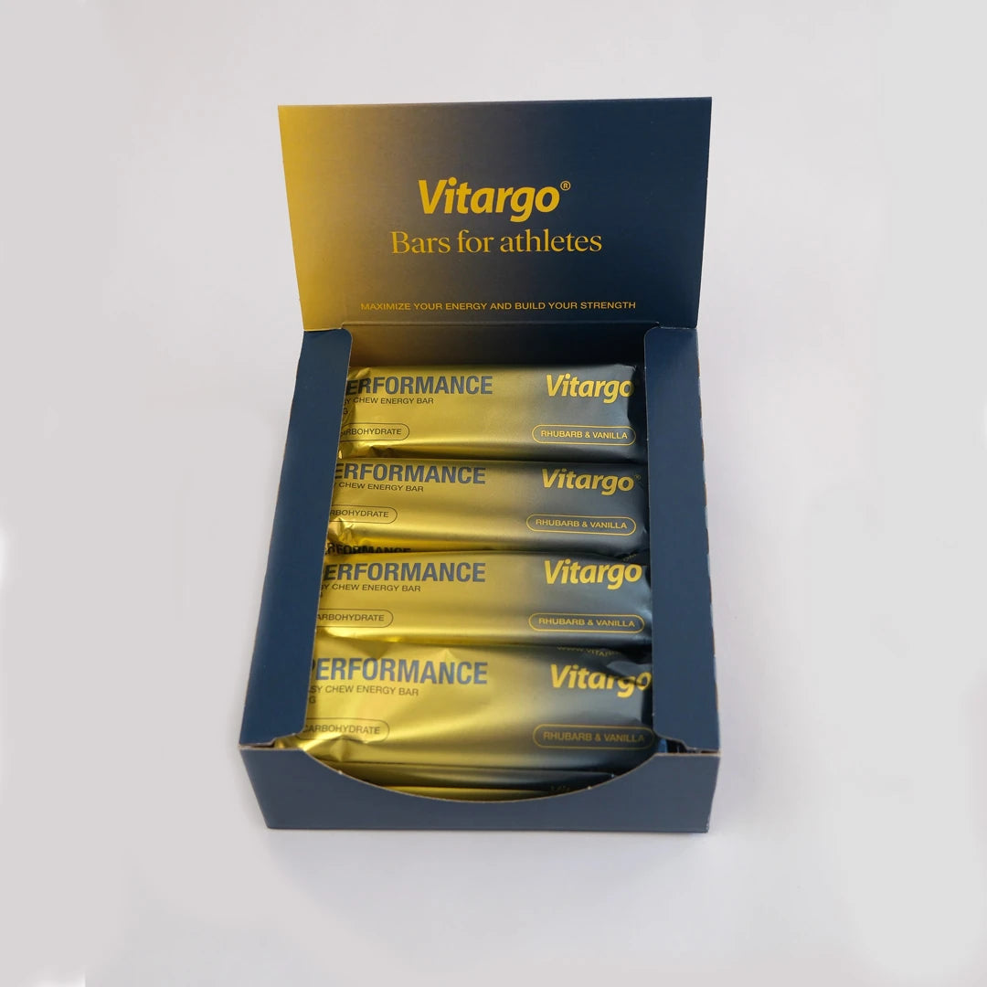 12 x Vitargo Performance Bar 55 g i en mörkblå låda fylld med guldiga energibars i smaken Rhubarb & Vanilla.